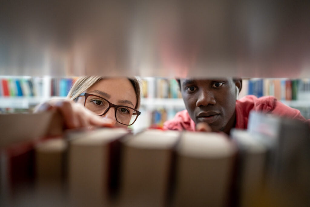 Closeup through bookshelf couple of students looking for right book in library to prepare for exam.